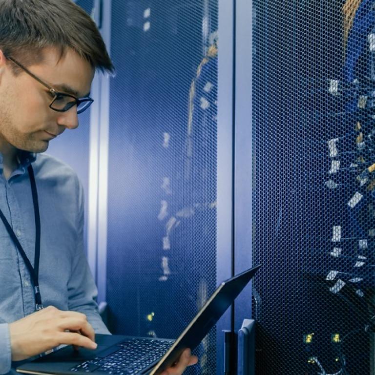 Picture of an IT guy looking at his laptop and standing in front of a rack with servers and cables 